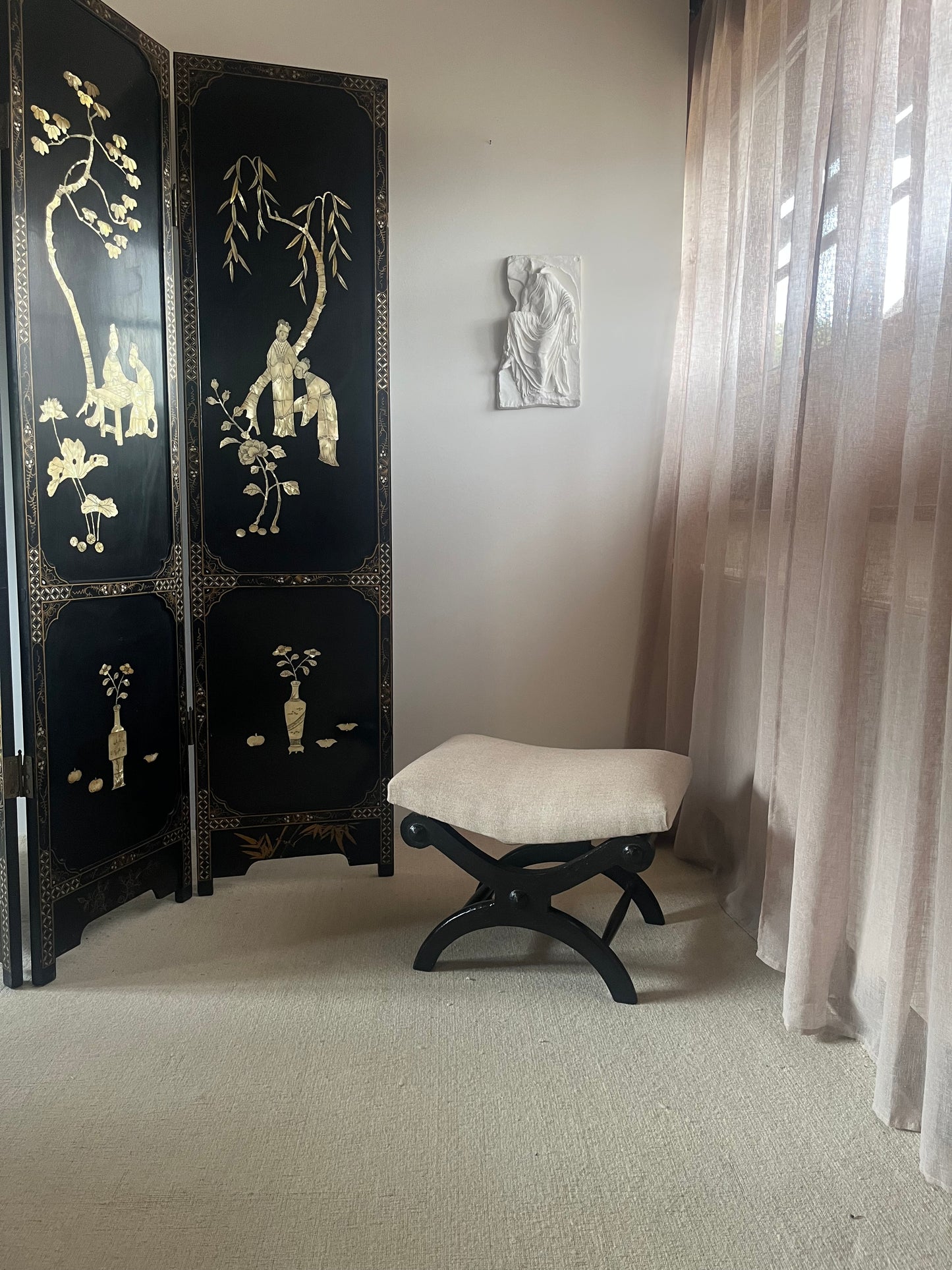 A black folding screen with gold decorative illustrations stands next to a small black stool with a cream cushion in a room with beige walls and sheer curtains.