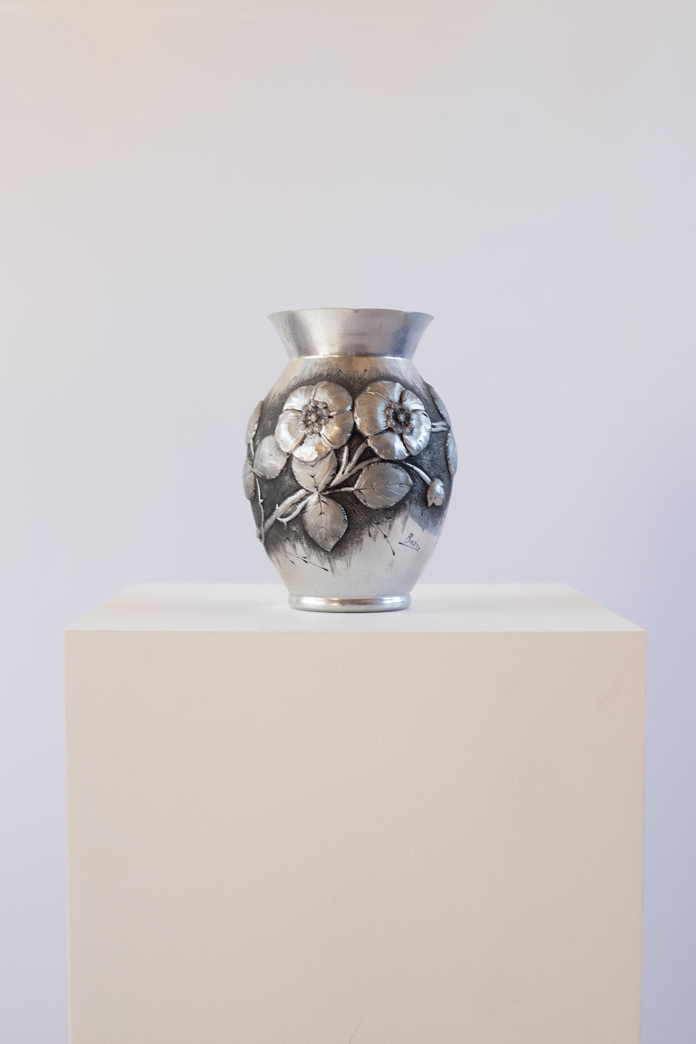Silver vase with floral design on a white pedestal against a white background