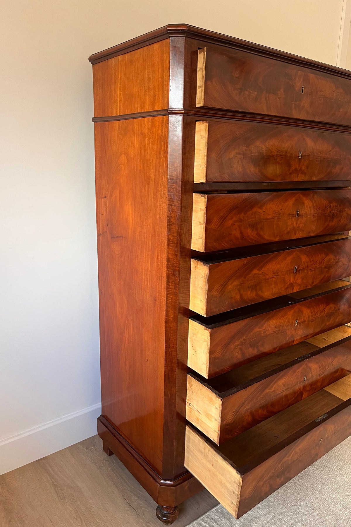 A tall, antique-style wooden chest of drawers with six drawers, shown in a home setting with one lower drawer pulled open. The chest is made of rich, warm brown wood with visible grain and light-colored wood edges.