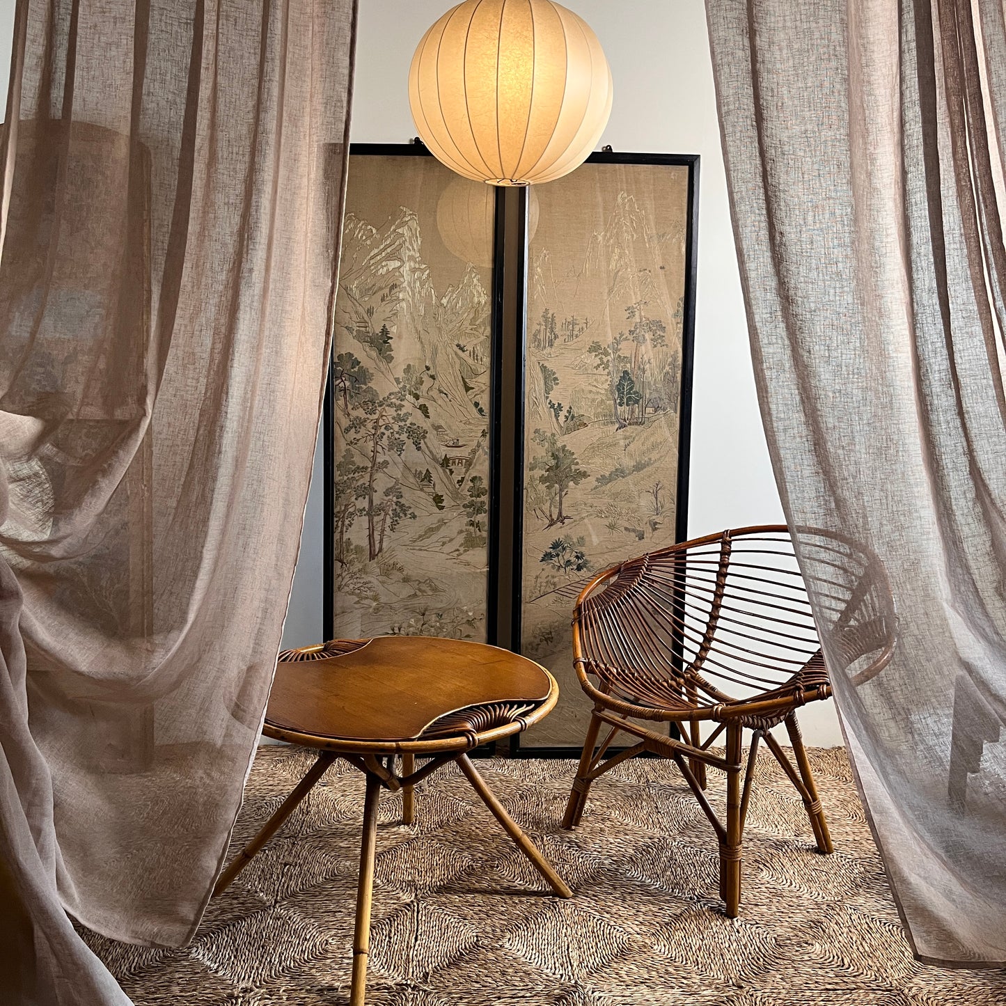 Wicker chair and table on a textured surface with a screen behind and brown drapes either side of them.