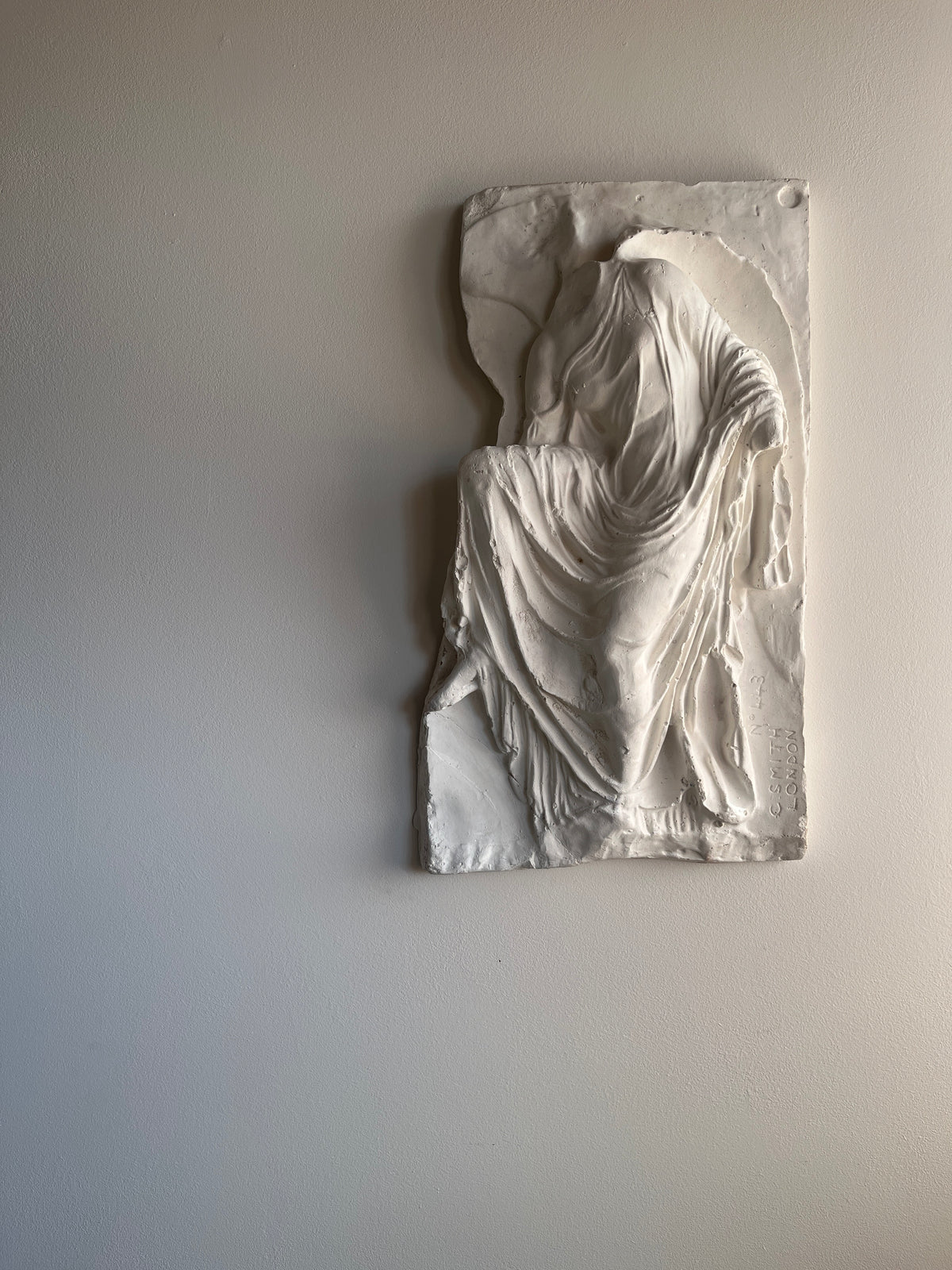 A white plaster relief sculpture of a draped, seated figure mounted on a light gray wall.