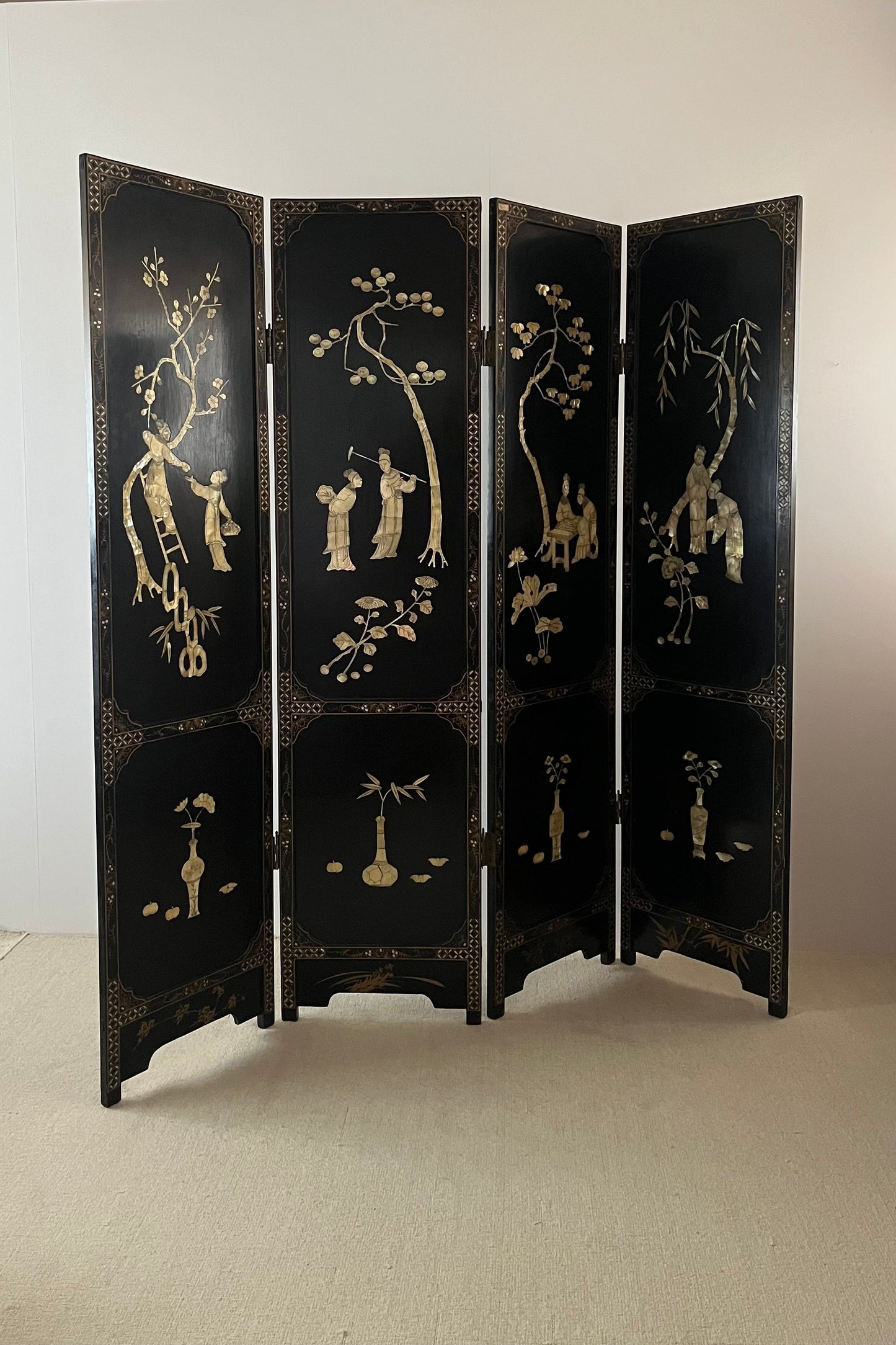 A four-panel folding screen with a glossy black lacquered finish and hand-painted gold chinoiserie-style illustrations of trees, figures, and decorative motifs, standing against a plain off-white wall.