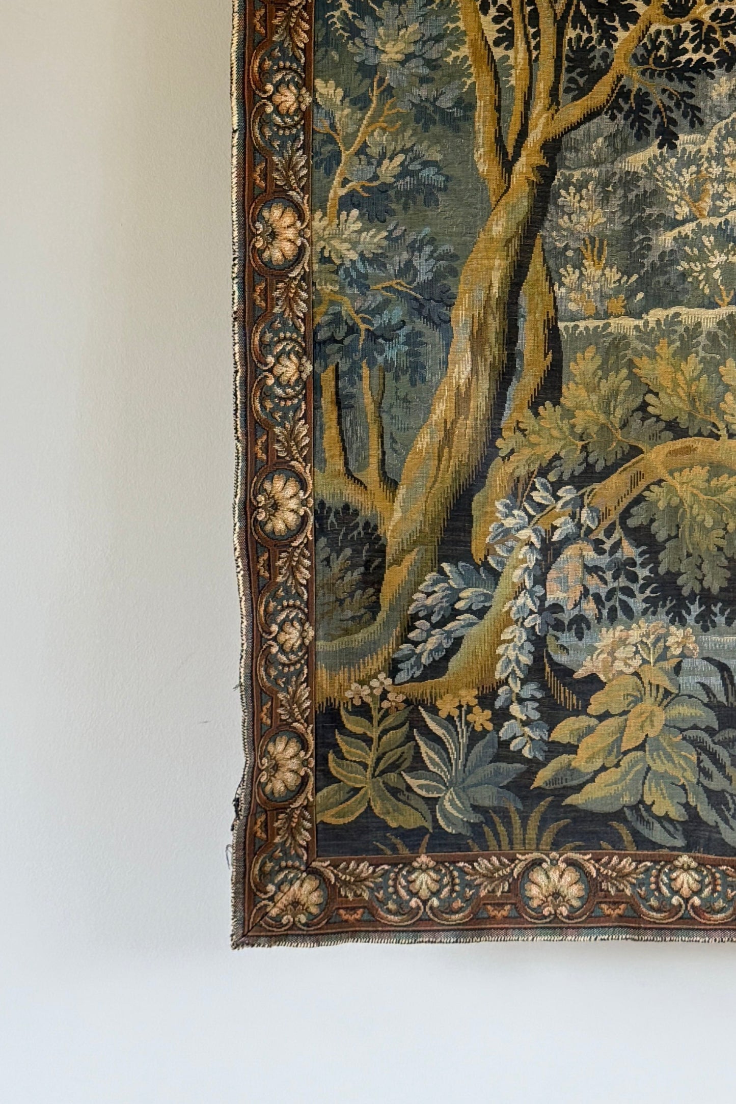 A square vintage tapestry featuring a detailed landscape scene with trees, foliage, and a deer, hung on a plain off-white wall.