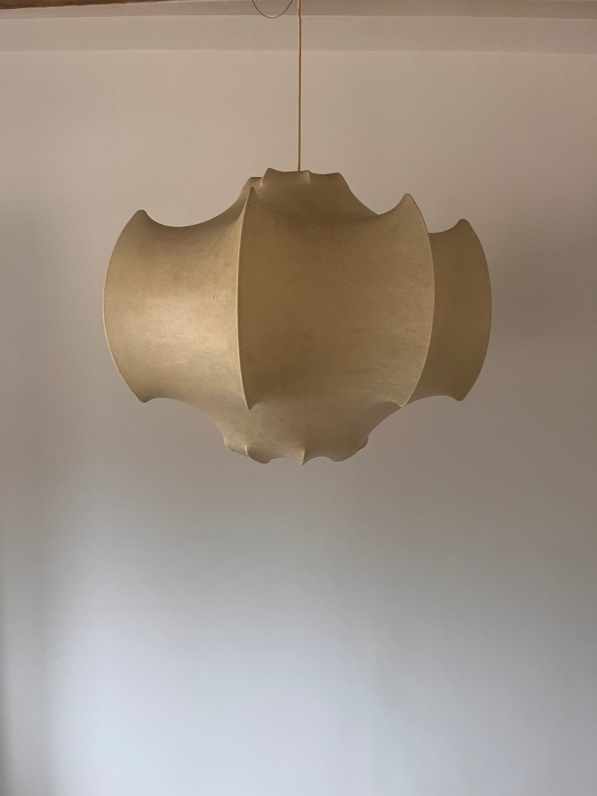 A modern, sculptural pendant light with a wavy, layered paper-like shade in a warm beige or light tan color, suspended from a thin cord against a plain off-white wall.