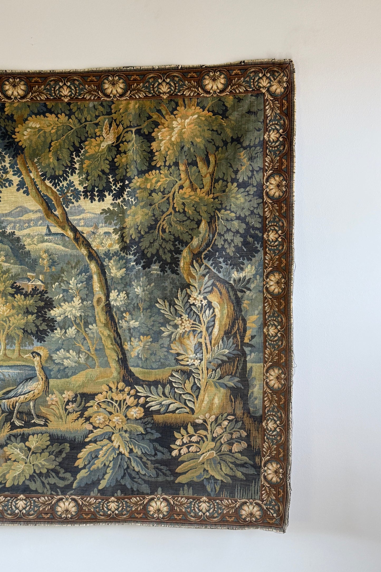 A vintage tapestry featuring a detailed pastoral landscape with trees, foliage, and a bird, shown hanging on a plain white wall.