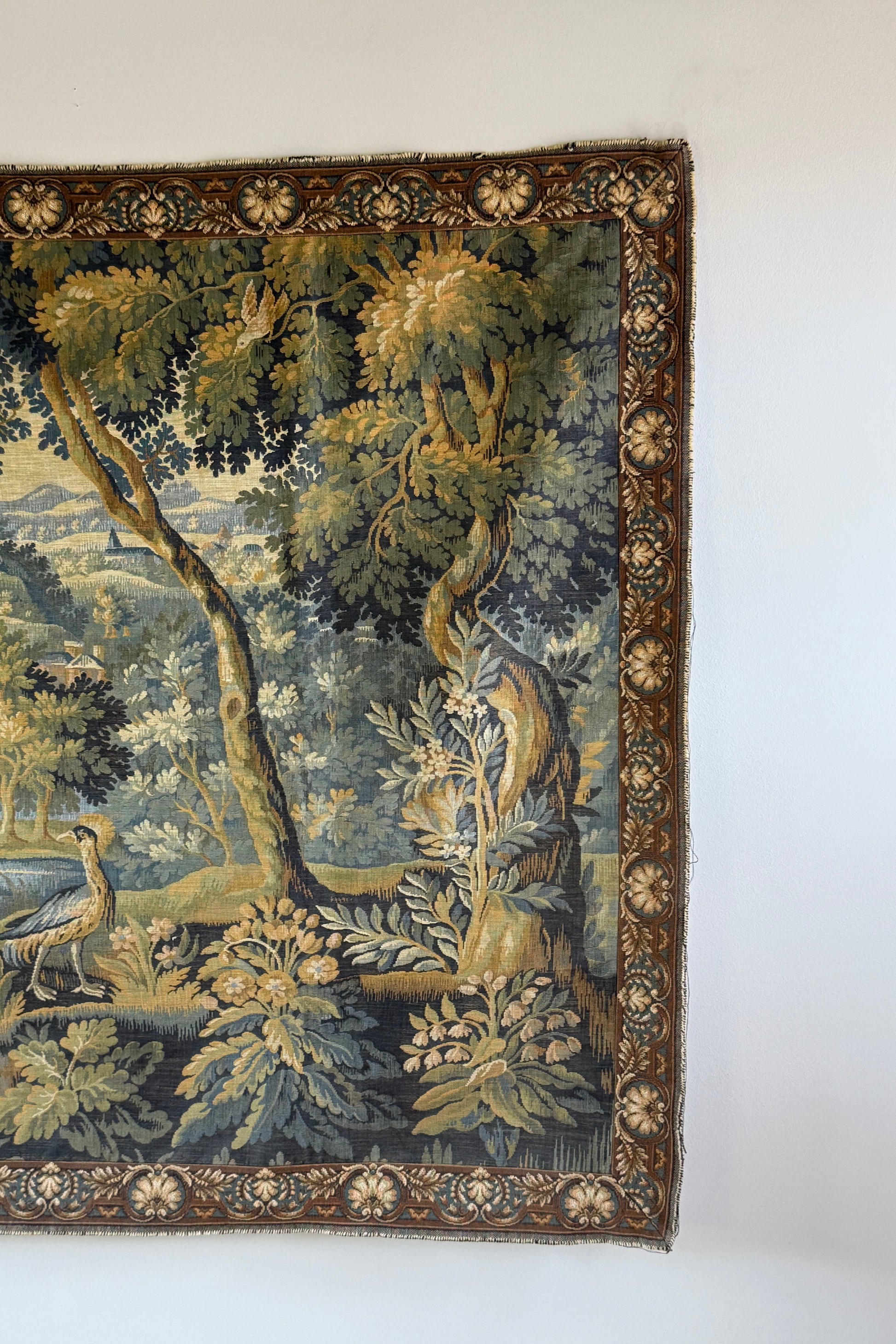 A vintage tapestry featuring a detailed pastoral landscape with trees, foliage, and a bird, shown hanging on a plain white wall.