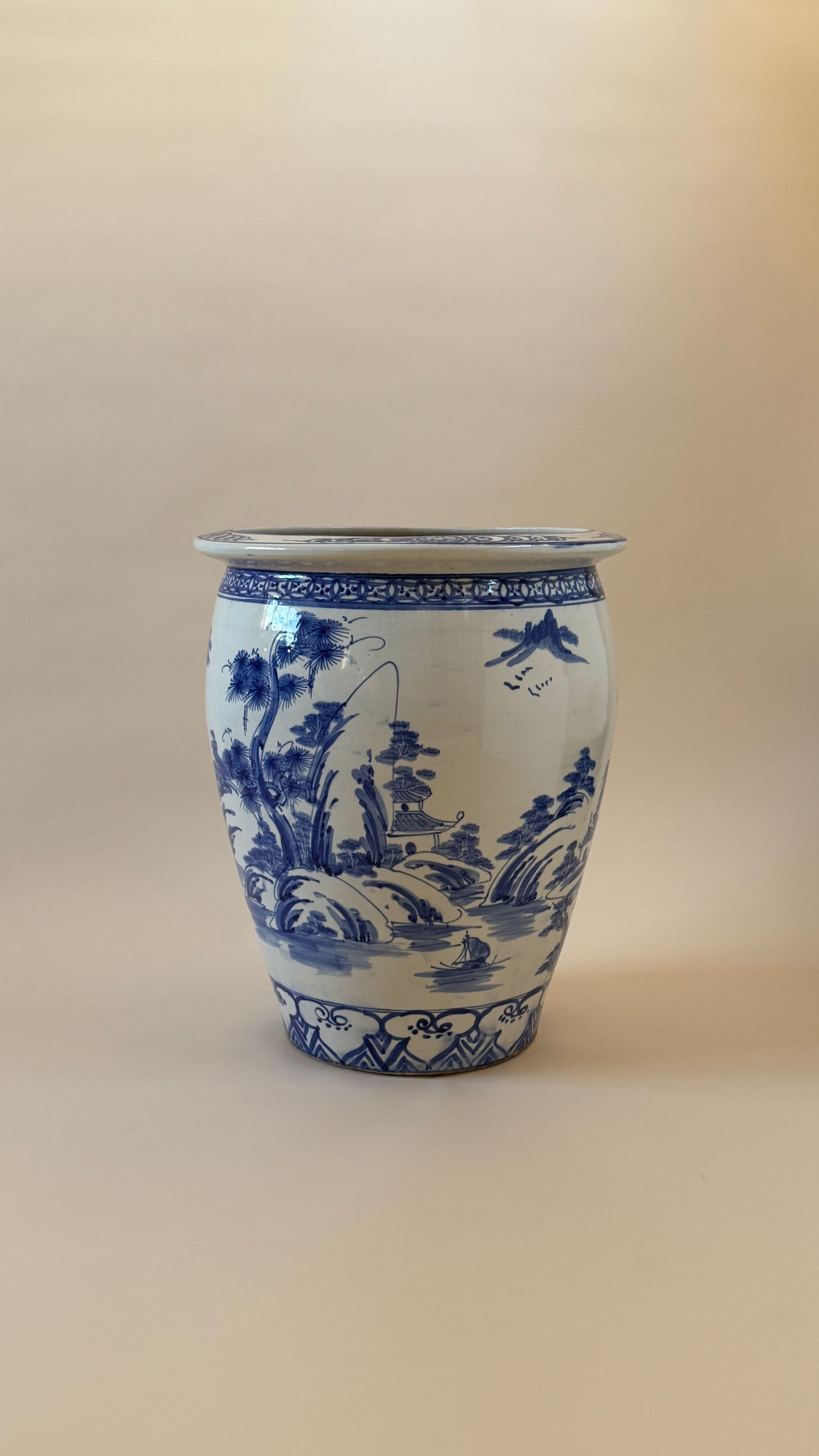 A classic blue and white ceramic planter pot featuring hand-painted traditional Chinese landscape scenes with mountains, trees, and birds, set against a plain beige background.