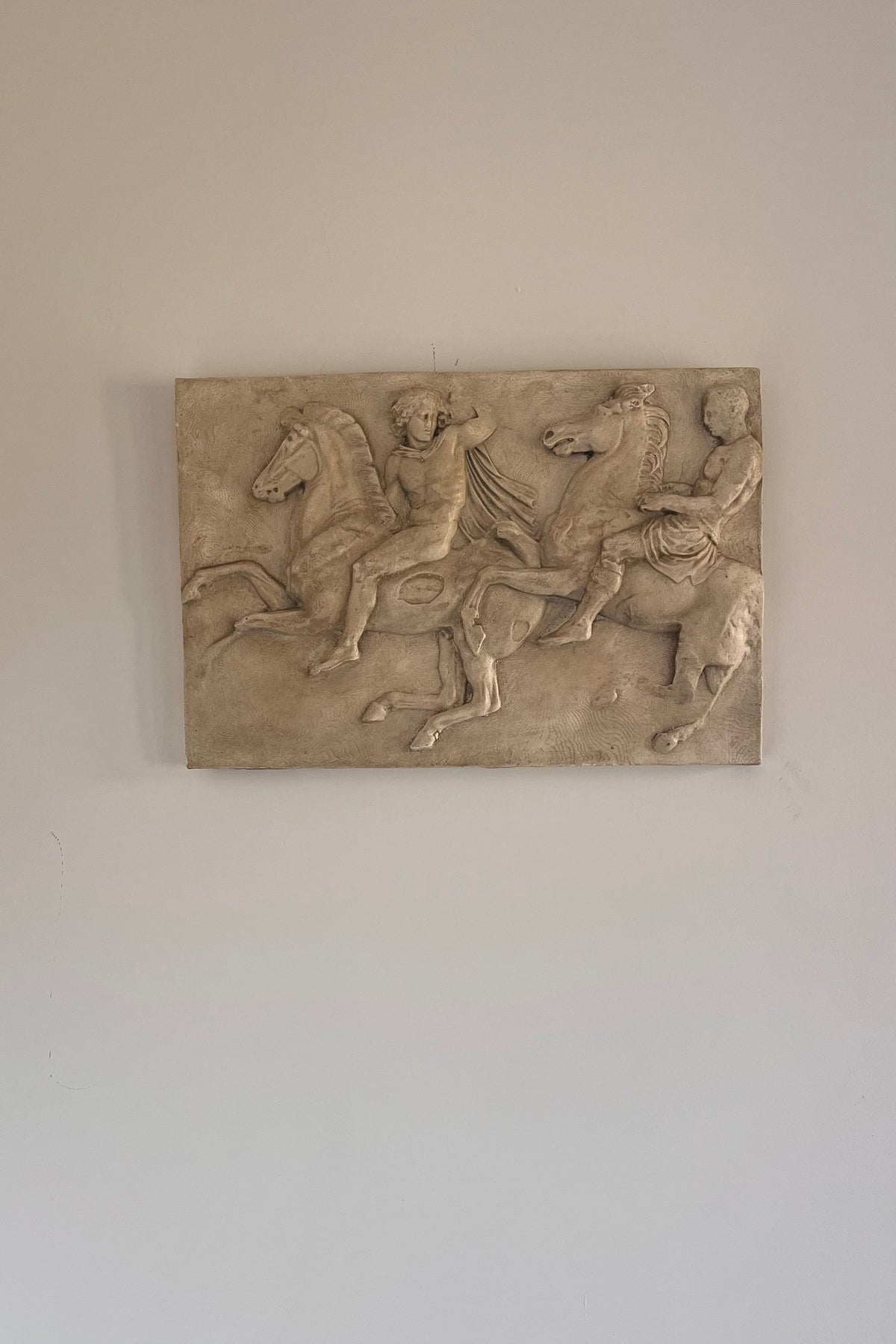 A rectangular stone relief sculpture depicting classical figures on horseback, mounted on a plain off-white wall.