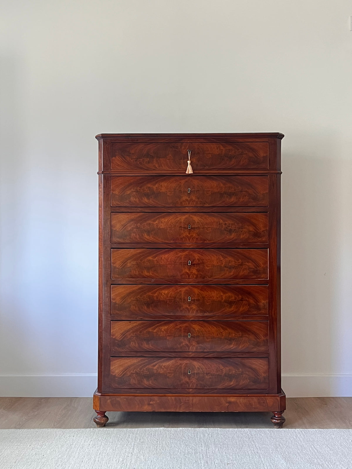 A tall antique wooden chest of drawers with seven drawers, featuring rich brown and amber walnut grain patterns, a small brass pull handle, and turned feet, set against a plain off-white wall and light wood floor.