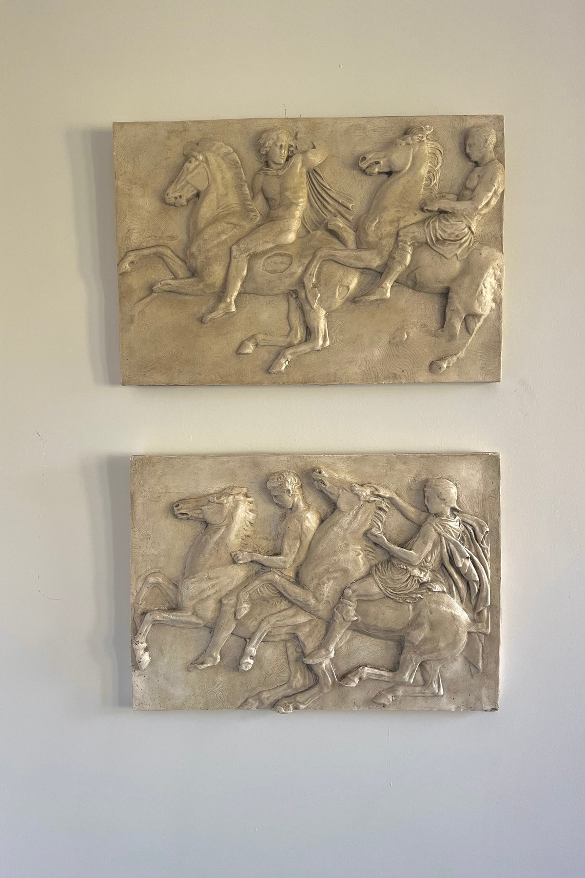 Two rectangular stone relief wall plaques depicting classical figures on horseback, mounted on a light gray wall.
