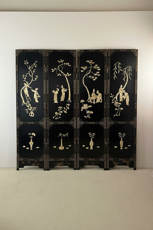 A six-panel folding lacquer screen with a glossy black finish and intricate gold hand-painted motifs of figures, trees, and floral designs, displayed against a plain white wall.