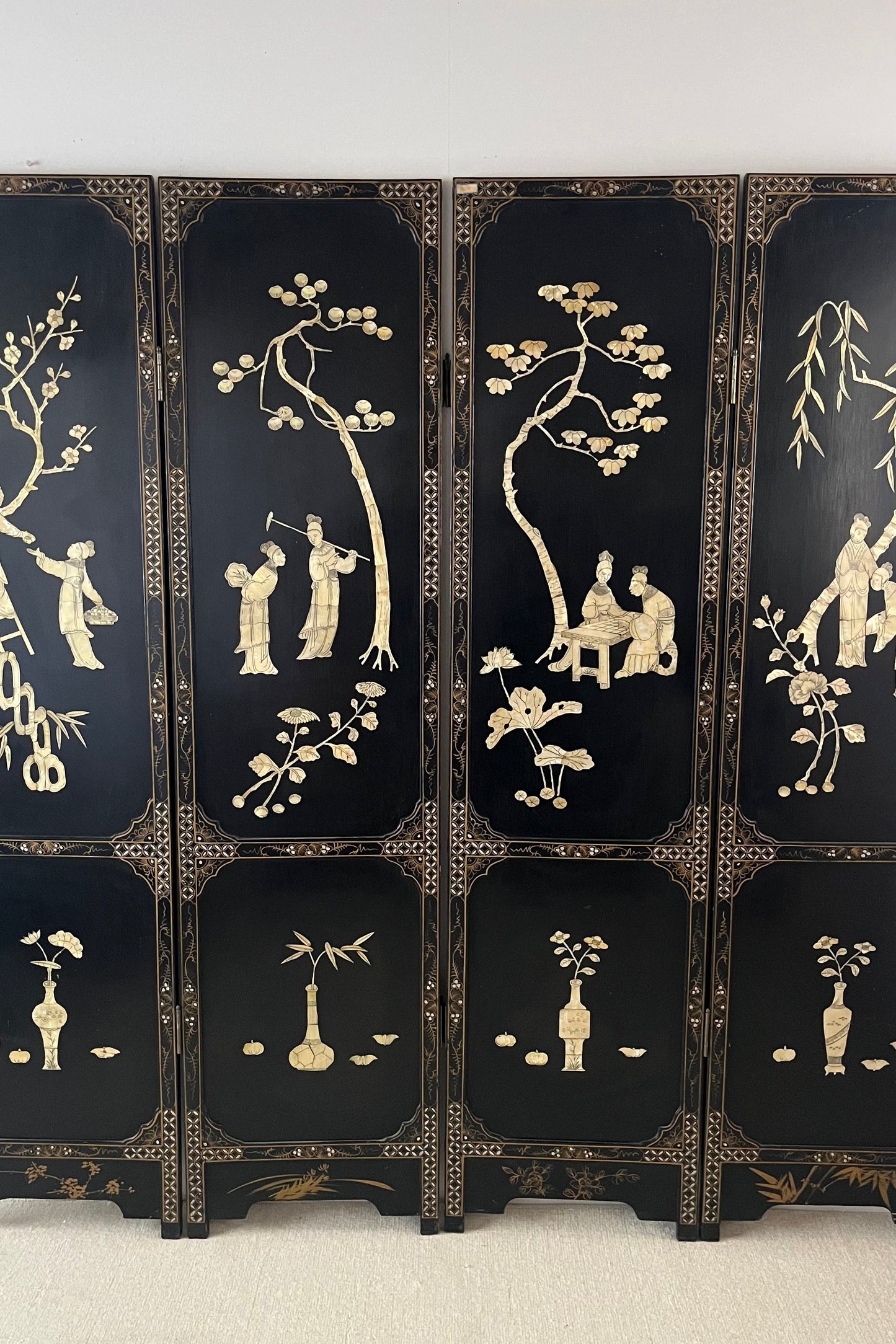 A multi-panel black lacquered folding screen with intricate gold and beige inlay depicting traditional figures, trees, and floral motifs, set against a plain light gray wall and light carpet.