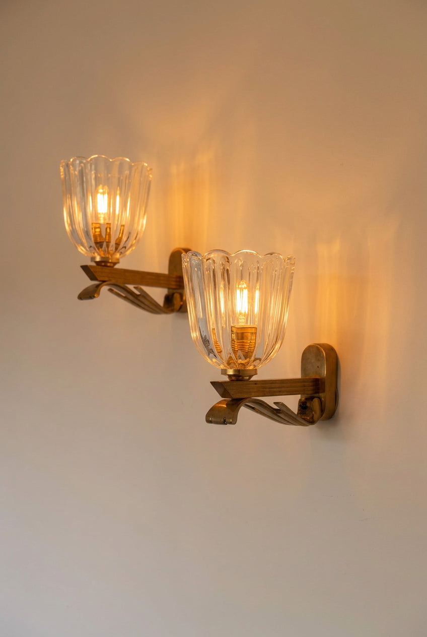 A two-light wall or ceiling mount fixture with clear ribbed glass shades and a warm brass or bronze metal frame, shown against a plain off-white wall.