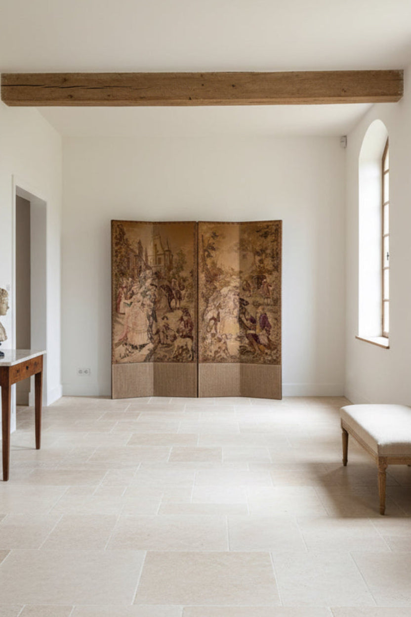 A two-panel folding screen with a vintage tapestry design, featuring a sepia-toned French village scene with figures and architecture, set against a white wall in a room with wooden ceiling beams.