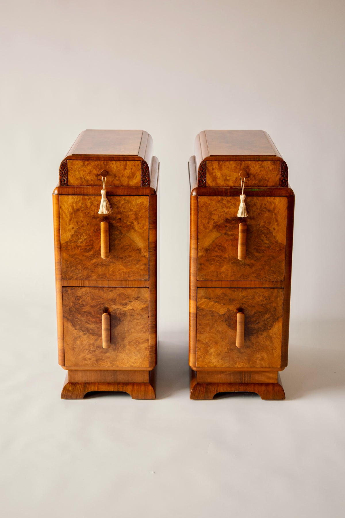 A pair of mid-century Art Deco wooden nightstands with burl wood grain, featuring two drawers each, brass pull handles, and tassel accents, set against a neutral light gray background.
