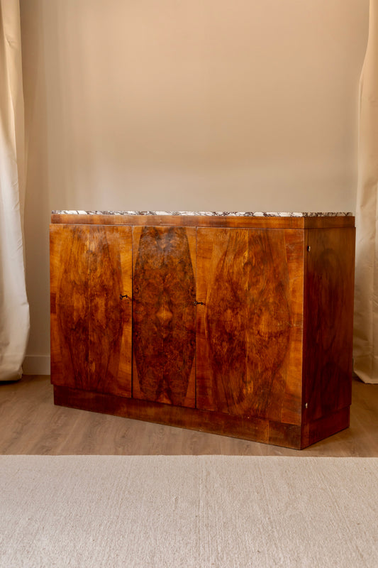 A vintage wooden sideboard cabinet with a highly figured burl wood grain finish and a polished marble top, set against a neutral beige wall and light wood floor.