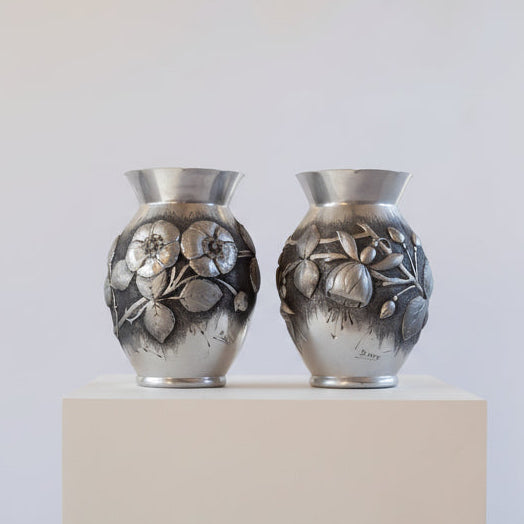 Two silver vases with floral designs on a white pedestal against a white background