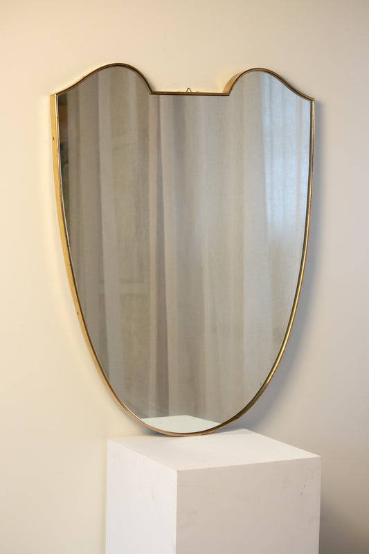 midcentury brass Italian
Shield-shaped mirror with brass frame on a white pedestal against a beige wall