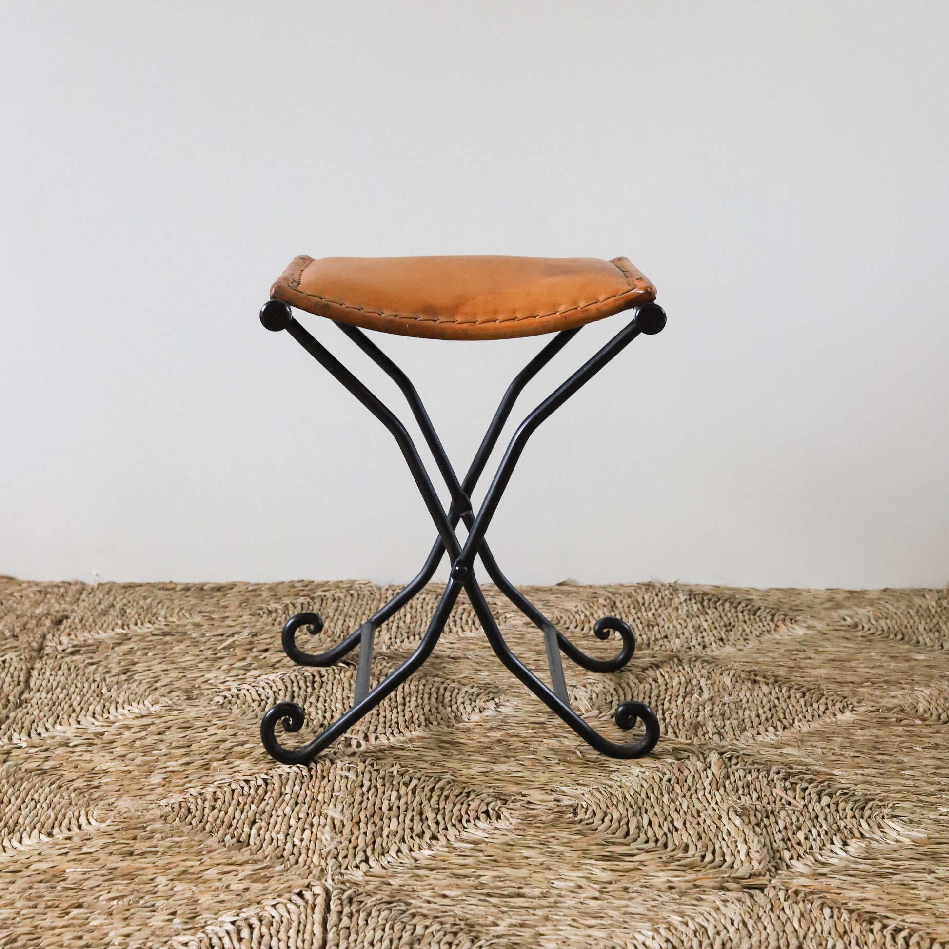 Small vintage stool with a brown leather seat and black metal frame on a textured surface.