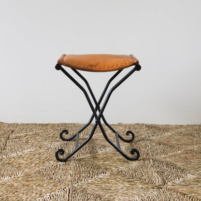 Small vintage stool with a brown leather seat and black metal frame on a textured surface.