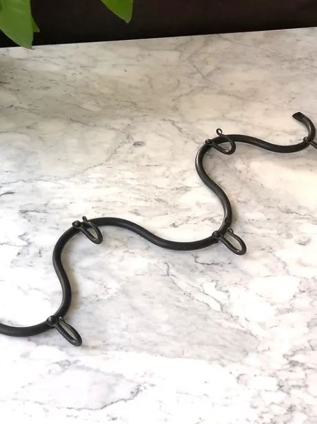 An iron Maud Vaughan Wave Hook laying flat on a marble table. Wiggle hooks