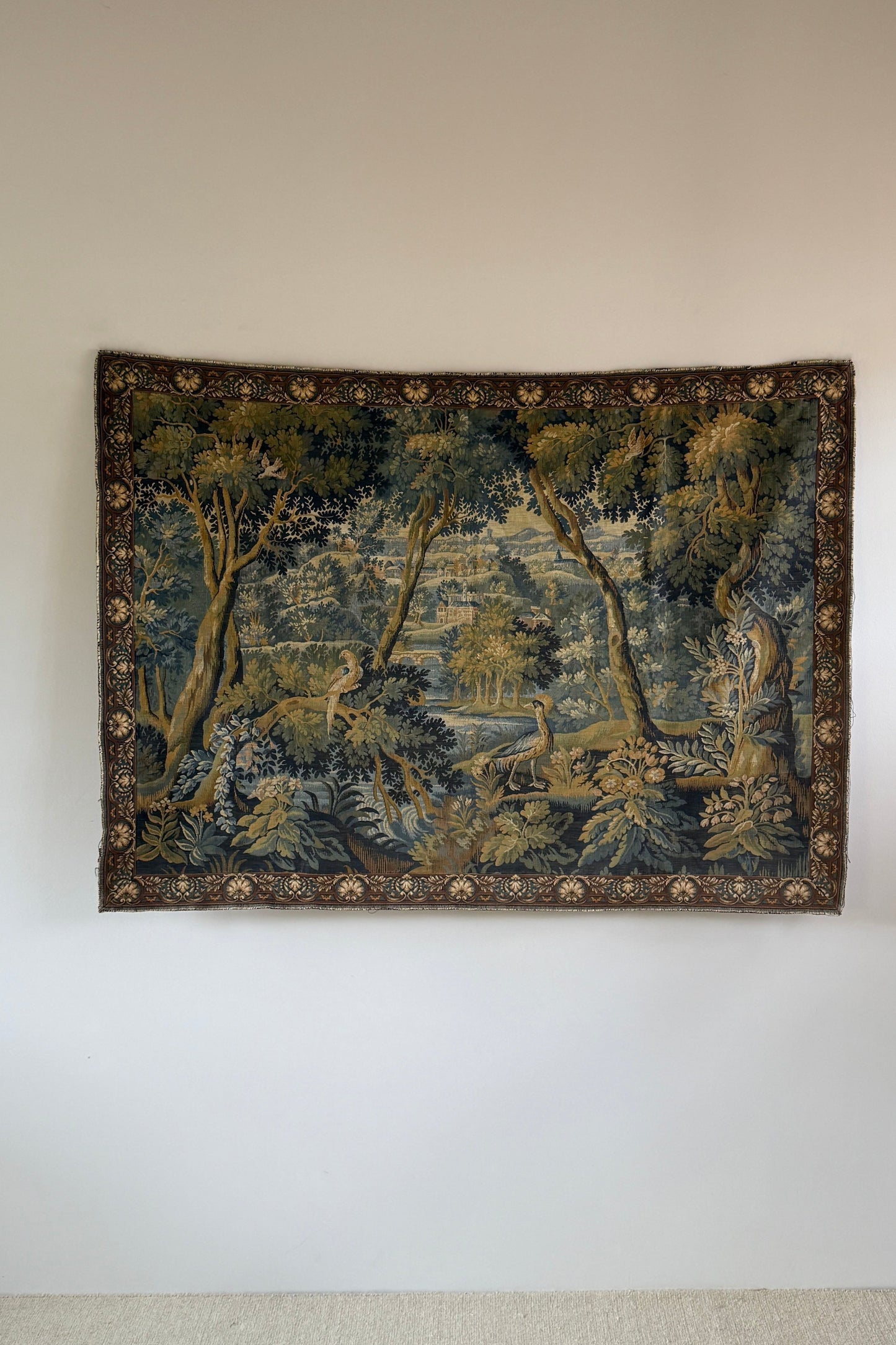 A rectangular vintage tapestry featuring a detailed landscape scene with trees, water, and figures, displayed on a plain white wall.