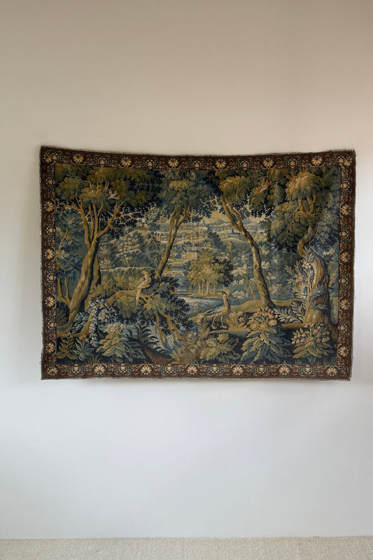 A rectangular vintage tapestry featuring a detailed landscape scene with trees, water, and figures, displayed on a plain white wall.